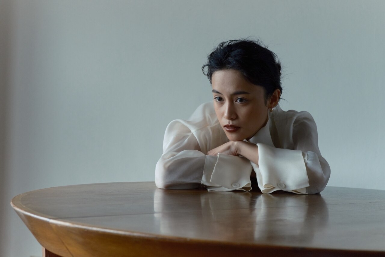 白いブラウスの女性が木のテーブルに頬杖をつく、落ち着いたアート的な雰囲気のポートレート